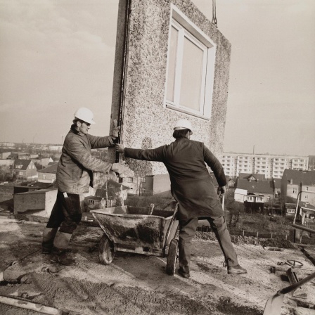 Einen Schwarz-Weiß-Aufnahme aus den 1970er Jahren zeigt Bauarbeiter beim Einfügen eines Teilstücks für ein Wohnhaus, das in Plattenbauweise errichtet wird.