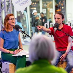 Annelie Malun sitzt links in einem blauen Oberteil vor einem Mikrofon auf einem Holzhocker und lacht. Christian Karn sitzt rechts von ihr auf einem Holzhocker vor einem Mikrofon und zeigt ins Publikum.