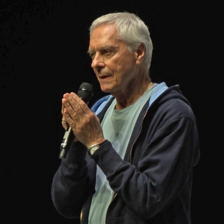 John Neumeier hält vor schwarzem Hintergrund sichtlich berührt das Mikro zwischen den Händen