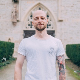 Christopher Schlicht steht vor einer Kirche, trägt ein weißes T-Shirt. Sein linker Arm ist tätowiert.