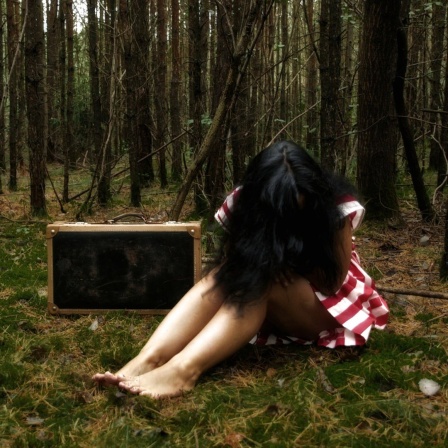Eine Frau sitzt neben einem Koffer im Wald, das Gesicht von ihren Haaren verdeckt.