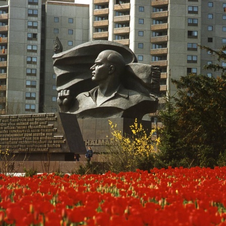 Das Ernst Thälmann Denkmal vor der Plattenbau-Kulisse in Berlin-Prenzlauer Berg. Im Vordergrund rote Blumen, im Hintergrund die Plattenbauten.
