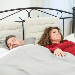 Eine Frau liegt wach in einem Bett, während ihr Mann bereits schläft