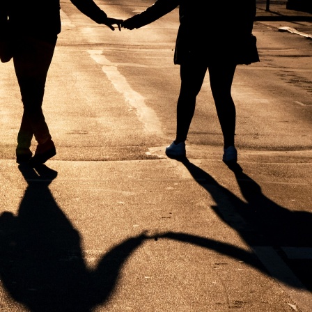 Zwei Menschen, die ihre Hände halten, gehen über einen abendsonnengeflutete Straße, sodass nur ihre Silhoutte und Schatten zu erkennen sind.