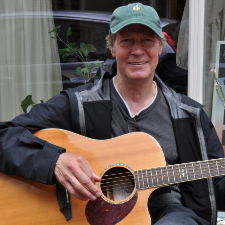 Der Musiker und Wirt Rainer Martens hält eine Gitarre in der Hand und lächelt in die Kamera.