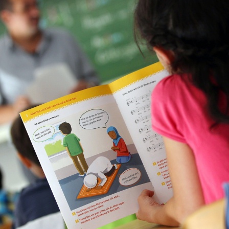 Symbolbild: Eine Mädchen liest während des islamischen Religionsunterrichts in einem Schulbuch. Im Hintergrund ist unscharf ein Lehrer zu sehen.